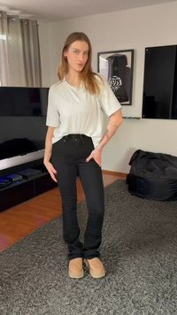 Ohlapna svetlo siva majica, črne jeans hlače z visokim pasom, rjavi copati, stoji na sivem preprogi z modernim dnevnim prostorom v ozadju.