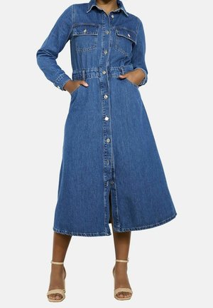 Femme en longue robe en jean bleu boutonnée avec poches et talons ouverts à bride cheville couleur beige, debout, les mains dans les poches.