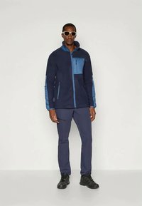 Giacca in pile blu navy con dettagli azzurri, chiusura a zip, colletto alto e tasca sul petto con zip. Abbinata a pantaloni blu navy e scarpe nere.