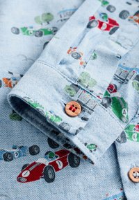 Tessuto per camicia in spina di pesce azzurro chiaro con motivi colorati di auto da corsa. Rifinito con un bottone in legno sul polsino.