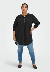 Tunika nera a maniche lunghe con placca di bottoni, abbinata a jeans chiari strappati e sandali neri a scivolo. Tessuto dalla texture liscia.