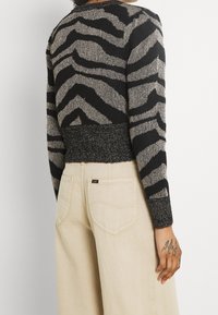 Pull en maille noir et argenté métallisé avec un motif à rayures zèbre, doté d'une ceinture côtelée et de manches longues. Accompagné d'un pantalon beige.