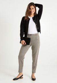 Femme souriante, portant un cardigan noir, un haut blanc, un pantalon à carreaux, des ballerines noires à talon, tenant une pochette noire, posant devant un fond blanc.