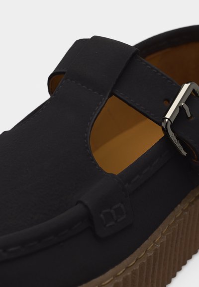 Μαύρο δερμάτινο σανδάλι με σχεδιασμό κοψίματος, ασημένια αγκράφα και χοντρή καφέ σόλα με αυλακώσεις, σε κοντινή λεπτομέρεια.