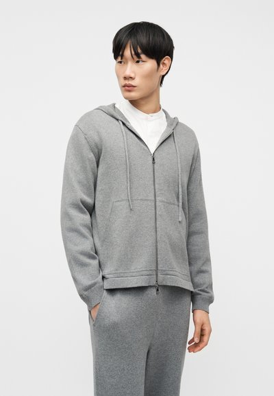 Νέος άντρας που φοράει γκρι φούτερ με φερμουάρ και ταιριαστό παντελόνι, κοιτάζει πλάγια με τα χέρια στις τσέπες, σε απλό φόντο.