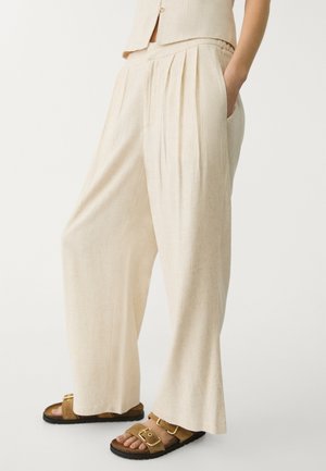Pantalon plissé beige en tissu léger avec poches latérales, associé à des sandales marron avec détails dorés et semelles noires.