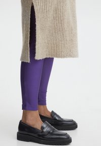 Maglione beige a costine con spacchi laterali abbinato a leggings viola aderenti e mocassini neri con suola in gomma spessa.