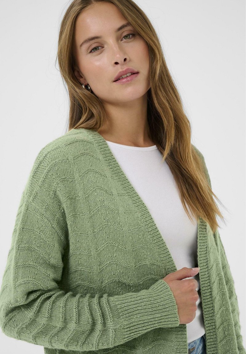 Lichtgroene cardigan met een textuur van golvende patronen, open voorkant en geribbelde manchetten. Draagt over een wit shirt, casual uitstraling.
