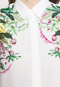 Blouse blanche ornée de broderies florales colorées en vert, rose et jaune. Comprend un col pointu et une fermeture à boutons.