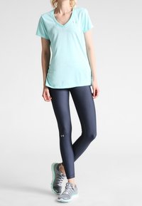 Kvinna som bär en ljusblå kortärmad V-ringad tröja, mörkblå leggings och grå sneakers, stående med korsade ben mot en vit bakgrund.