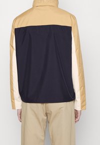 Person som bär en färgblock-jacka med marinblå rygg, beige axel och ärmar, samt beige sidostråk, i kombination med beige byxor.