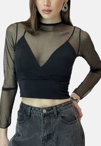 Zwart cropped top met lange doorschijnende mouwen van mesh, diepe V-hals en aangesloten silhouet. Gecombineerd met zwarte hoge taille spijkerbroek.