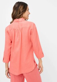 Coral linen shirt with long sleeves, featuring a buttoned collar, back pleating, and side slits for ease of movement.