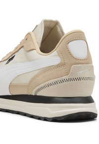 Zapatilla en beige y blanca con parte superior de ante y cuero, forma redondeada, talón texturizado y suela negra contrastante. Presenta detalles de costura.