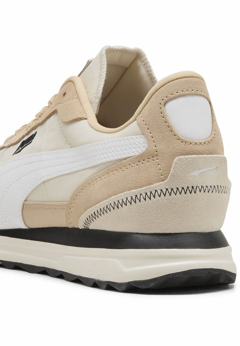 Zapatilla en beige y blanca con parte superior de ante y cuero, forma redondeada, talón texturizado y suela negra contrastante. Presenta detalles de costura.