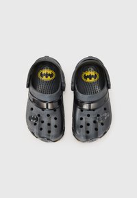 Black Crocs with ventilation holes, featuring a yellow Batman logo on the insole and a black bat symbol on the side. Soft, lightweight material.