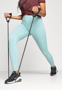 Svetlomodré legíny s vysokým pásom, čierne fitness pásy v rukách, skombinované s čiernymi športovými topánkami a tmavo vínovým tričkom s uzlom.