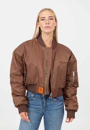 Blonde vrouw met een bruin bomberjack met geribbelde manchetten en tailleband, blauwe spijkerbroek, staand tegen een witte achtergrond.