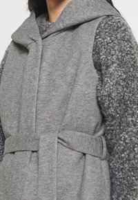 Cappotto grigio senza maniche con cappuccio, realizzato in tessuto testurizzato. Presenta una fascia in vita e un morbido maglione grigio peloso sotto.