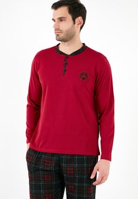 Rød langærmet skjorte med rund krave og fire knapper, med et sort logo patch, parret med sort og rød ternet bukser.
