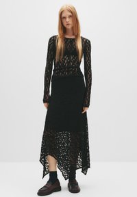 Top in pizzo nero con maniche lunghe, abbinato a una gonna in pizzo nero con taglio alto-basso. Caratterizzato da trame stratificate e un motivo floreale. Mocassini marroni e calze.