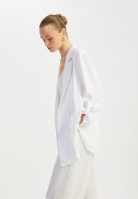 Blazer blanco de doble botonadura con solapas muesca, ajuste ligeramente holgado y mangas arremangadas, combinado con una parte superior de color claro y pantalones.