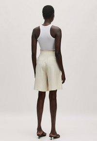 Beige linnen shorts met een tailormade pasvorm, een middelhoge tailleband en zakken; gecombineerd met een fitting wit tanktop. Soepele textuur en strakke lijnen.