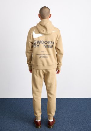 Persona in piedi di spalle che indossa una felpa con cappuccio e pantaloni beige Nike, con il logo Nike bianco e testo sul retro della felpa, scarpe marroni.