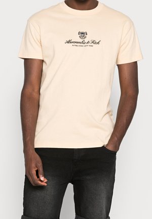 Beiges Baumwoll-T-Shirt mit Rundhalsausschnitt, das in der Mitte ein schwarzes gedrucktes Logo mit Text zeigt. Gerade geschnitten, kurze Ärmel und Standard-Passform.