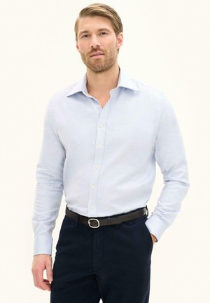 Mann mit hellbraunen Haaren und Bart trägt ein hellblaues Hemd, eingesteckt in dunkle Hose mit schwarzem Gürtel, steht vor schlichtem Hintergrund.