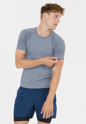 Ung mand i blå atletisk skjorte og shorts, der justerer sin ærme og kigger til siden mod en ensartet hvid baggrund.