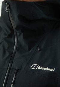 Jachetă impermeabilă neagră cu guler înalt, dotată cu un fermoar frontal și sireturi ajustabile; include un logo alb "berghaus" pe piept.