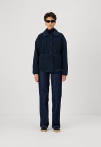 Giacca in fleece testurizzata blu navy con chiusura a bottoni, due tasche frontali, abbinata a pantaloni in denim scuro e sneakers. Occhiali da sole scuri indossati.