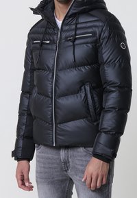 Homme portant une doudoune noire avec capuche, fermeture éclair frontale, poches poitrine zippées, et jean gris clair, debout devant un fond uni.