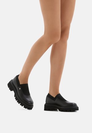 Stivali alla caviglia in pelle nera con parte superiore elastica simile a un calzino, suola in gomma spessa e texture ribattuta all'apertura. Design massiccio e moderno.