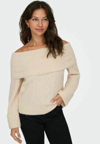 Beżowy sweter z odkrytymi ramionami o strukturze prążkowanej, z wzorami w splocie warkoczowym. Posiada szerokie mankiety i luźny krój. Zestawiony z czarnymi spodniami.