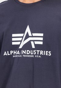 Laivastonsininen puuvillapaita, jossa on valkoinen graafinen logo, johon on lisätty tyyliteltyjä kirjaimia ja raitoja. Sisältää tekstin "Alpha Industries" ja sijaintitietoa.