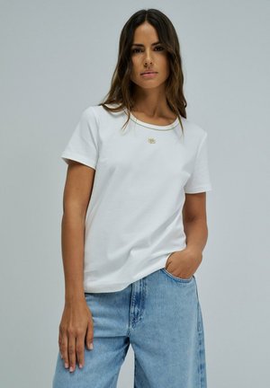 Mujer con cabello largo y oscuro, vestida con una camiseta blanca de manga corta y vaqueros vaqueros azul claro, de pie frente a un fondo liso de color gris claro.