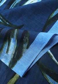 Tessuto blu con motivi di foglie verdi, superficie testurizzata e bordi arrotolati, che evidenziano una combinazione di tonalità blu scuro e chiaro.