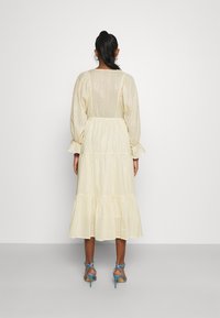 Maxi dress giallo chiaro con lunghe maniche a sbuffo, orlo a più strati e righe verticali. Abbinato a tacchi a strappo blu su uno sfondo grigio.