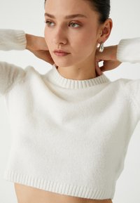 Witte cropped sweater van zachte, gestructureerde stof met een ribbelachtige halslijn en boorden. Het model heeft haar handen achter haar hoofd, waarmee de ontwerpelementen worden getoond.