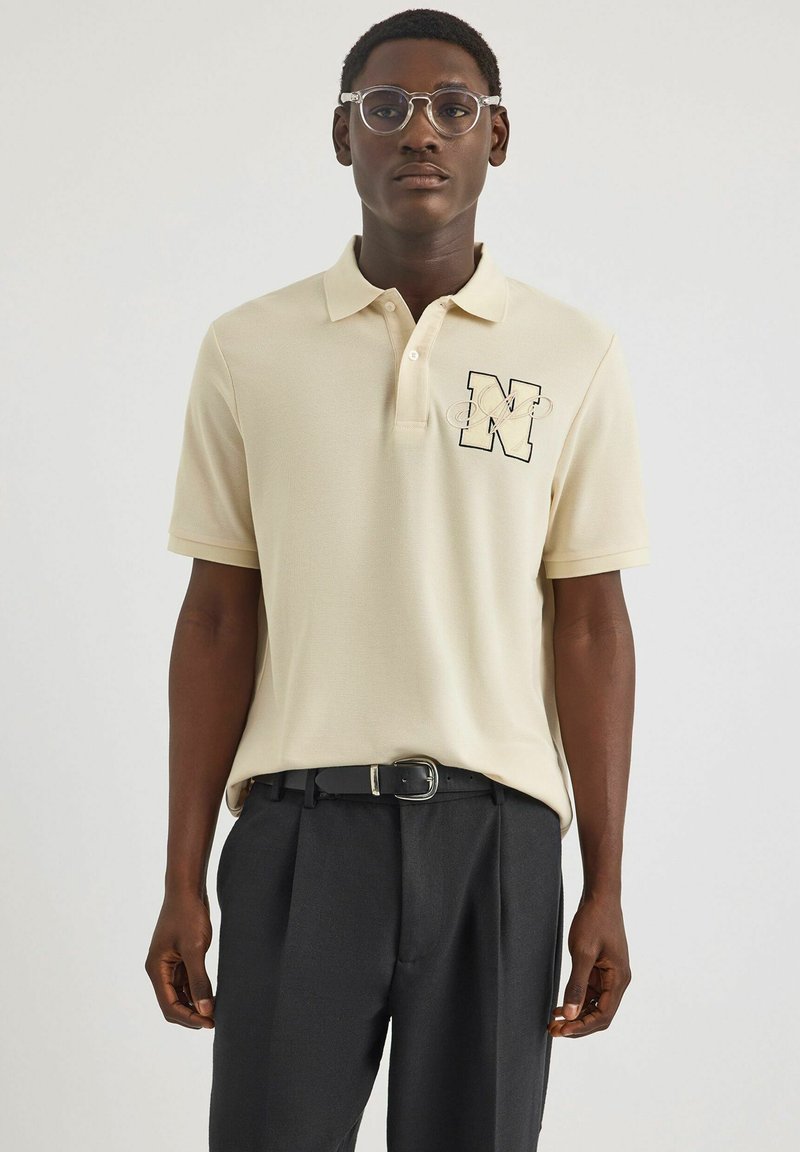 Beige Polohemd mit einem schwarz umrandeten Buchstaben "N" auf der linken Brust, kombiniert mit dunklen Hosen und einem schwarzen Gürtel. Strukturiertes Gewebe, klassische Passform.