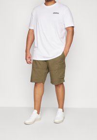 Vit t-shirt med kort ärm och texten "ORIGINALS", tillsammans med khaki cargo-shorts med fickor, och vita sneakers.