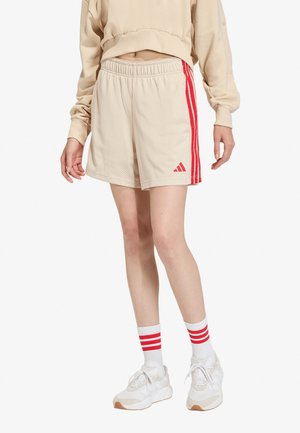 Persona que lleva puestos pantalones cortos deportivos beige de malla con rayas rojas y logo, zapatillas blancas y calcetines blancos con rayas rojas.