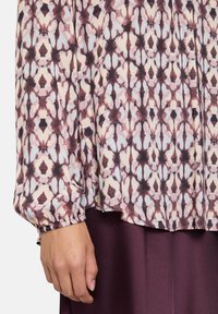 Blusa stampata con un motivo rosa, beige e marrone scuro. Maniche lunghe con polsini arricciati. Tessuto morbido e leggero che scivola con facilità.