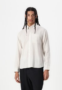 Chemise à manches longues beige clair à boutons avec un motif à carreaux, poche avant et tissu texturé. Portée avec un pantalon foncé.