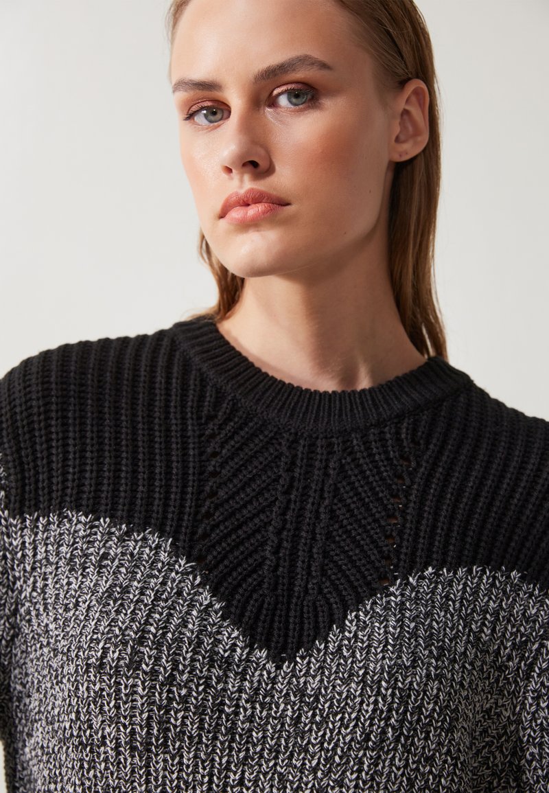 Sweter robiony na drutach z wzorem zygzakowym w czerni i szarości, z teksturowanym wykończeniem i okrągłym dekoltem. Zbliżenie, przycięty widok.