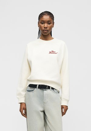 Jauna sieviete ar bizēm, tērpusies baltā New Balance džemperī un gaiši zilās džinsās ar melnu jostu, stāvot pret vienkrāsainu fonu.