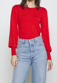 Rotes langärmliges Strickoberteil mit rundem Ausschnitt und Puffärmeln, kombiniert mit hellblauen hochtaillierten Jeans.