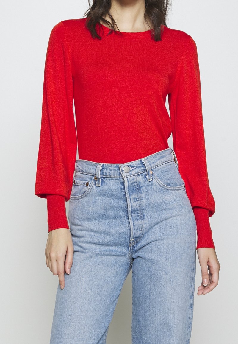 Rotes langärmliges Strickoberteil mit rundem Ausschnitt und Puffärmeln, kombiniert mit hellblauen hochtaillierten Jeans.